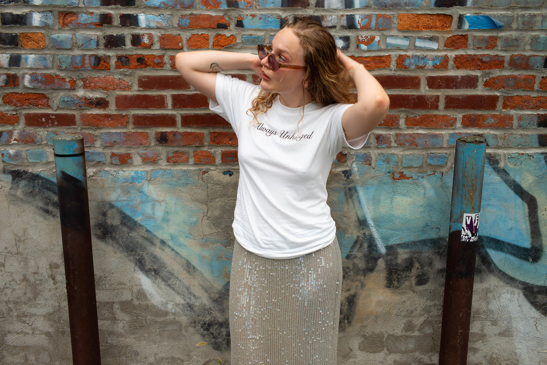 A woman with glasses posing in front of a brick and graffiti wall, wearing a white graphic t-shirt that says "Always Unhinged" and a sparkly beige maxi skirt.
