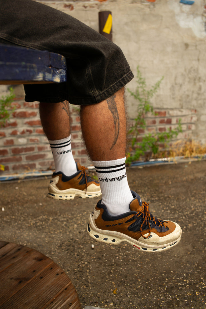 A person sitting on a bench wearing brown and beige chunky sneakers with white socks featuring the "Unhinged" brand logo, set against an urban brick wall background.