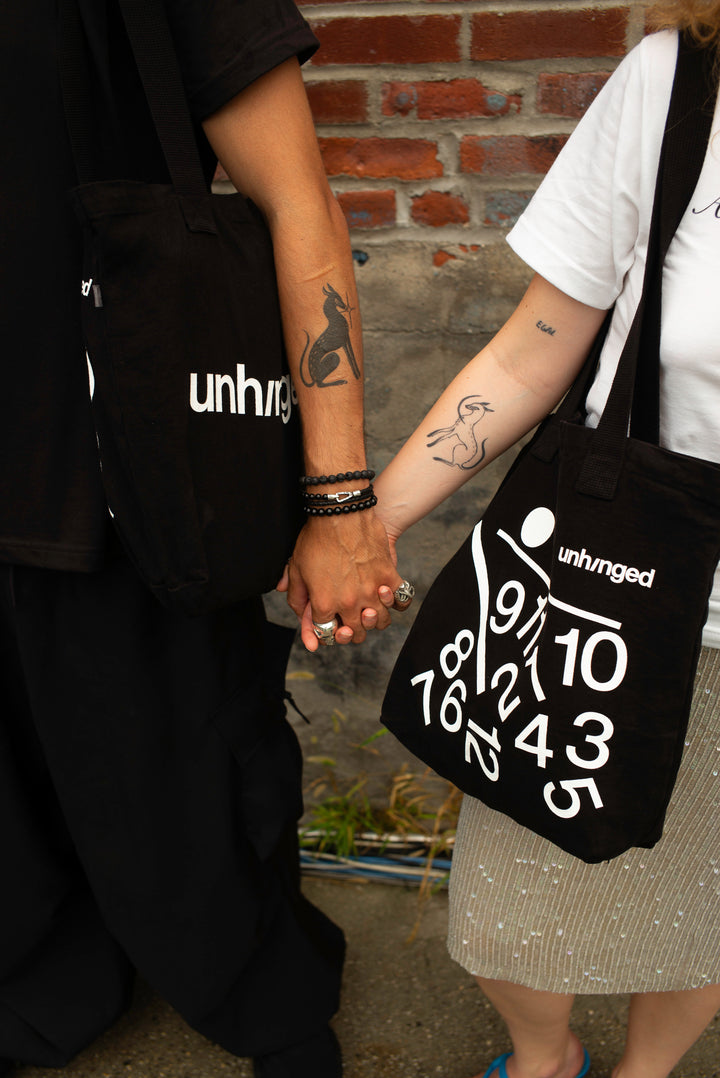 Two people holding hands while wearing matching black tote bags with white "Unhinged" branding and graphic prints, set against a brick wall background.