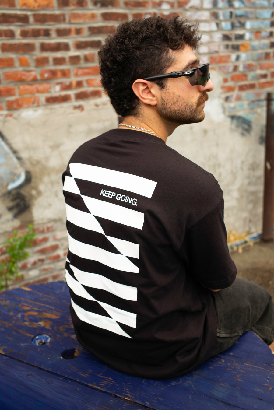 A man wearing a black t-shirt with a bold white striped 'KEEP GOING.' graphic on the back, sitting on a weathered blue bench against a brick wall.